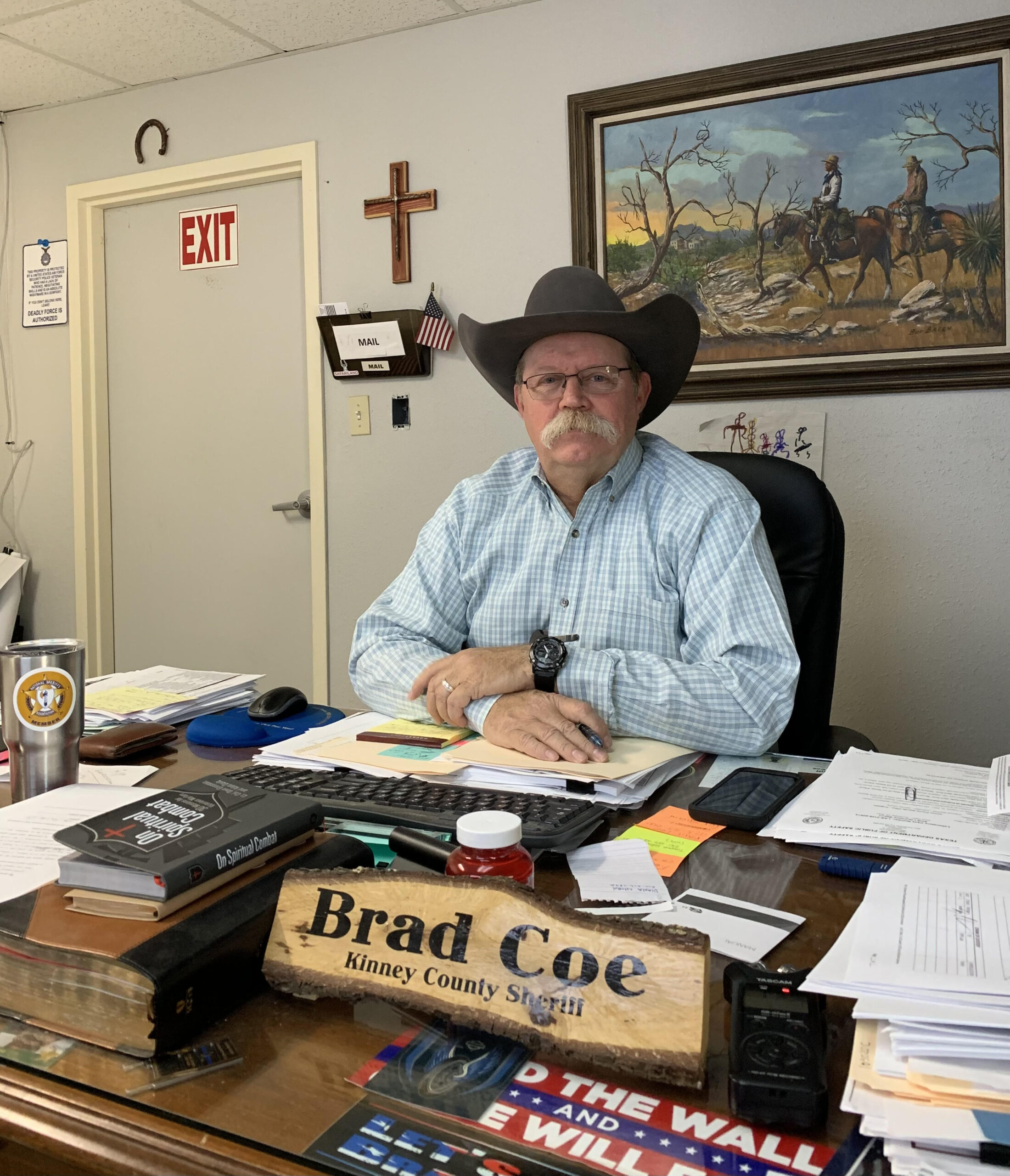 Sheriff Brad Coe sits behind a large nameplate at his desk in his office. He wears a cowboy hat and there is a cross on his wall.